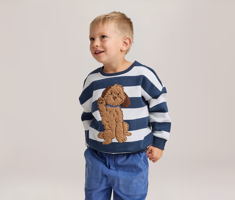 Ein kleiner Junge steht lächelnd und blickt nach oben. Er trägt ein weiß-blau gestreiftes Sweatshirt mit einer braunen Hundeapplikation und eine blaue Cordhose.