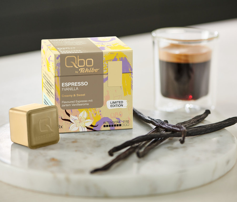 Eine Qbo Flavoured Espresso Vanilla - 8 Kapseln Packung, Kapsel, Espresso und Vanilleschoten auf einem Marmortablett neben einem Glas.