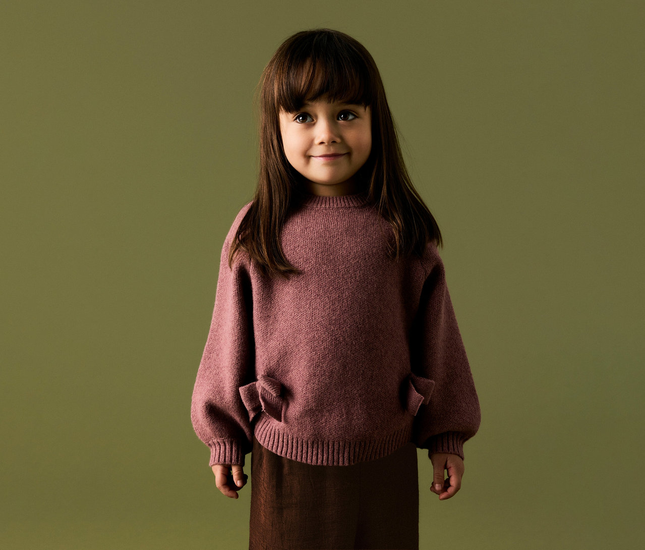 Junges Kind mit langen dunklen Haaren und Pony, lächelnd, trägt einen altrosafarbenen Strickpullover mit Puffärmeln und Schleifendetails sowie eine dunkelbraune Hose vor olivgrünem Hintergrund.