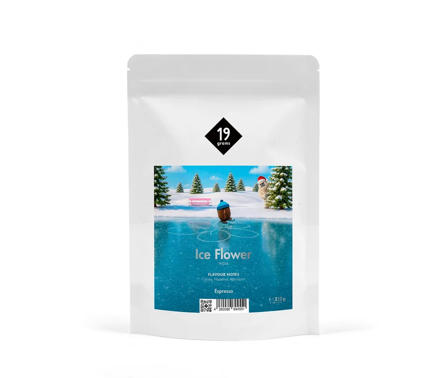 Weiße Kaffeeverpackung mit dem Logo „19 grams“ und einer Illustration einer Winterlandschaft mit einer Figur, die auf einer Eisfläche Schlittschuh läuft, umgeben von verschneiten Bäumen und einer Bank. Darunter steht „Ice Flower“, „INDIA“, „FLAVOUR NOTES Cacao, Hazelnut, Marzipan“, „Espresso“ und „250g“.