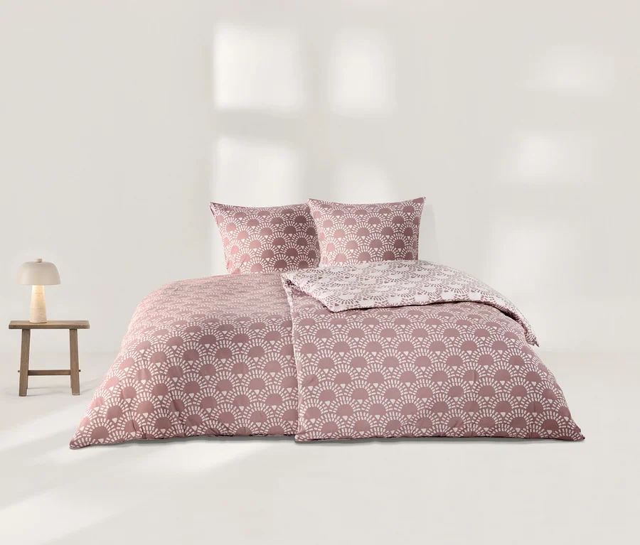 Rosa Jersey-Bettwäsche mit weißem Fächermuster, bestehend aus zwei Kissen und einem Bettbezug, dessen eine Seite ein helleres Muster zeigt. Links steht ein Holzschemel mit einer beigen Lampe.