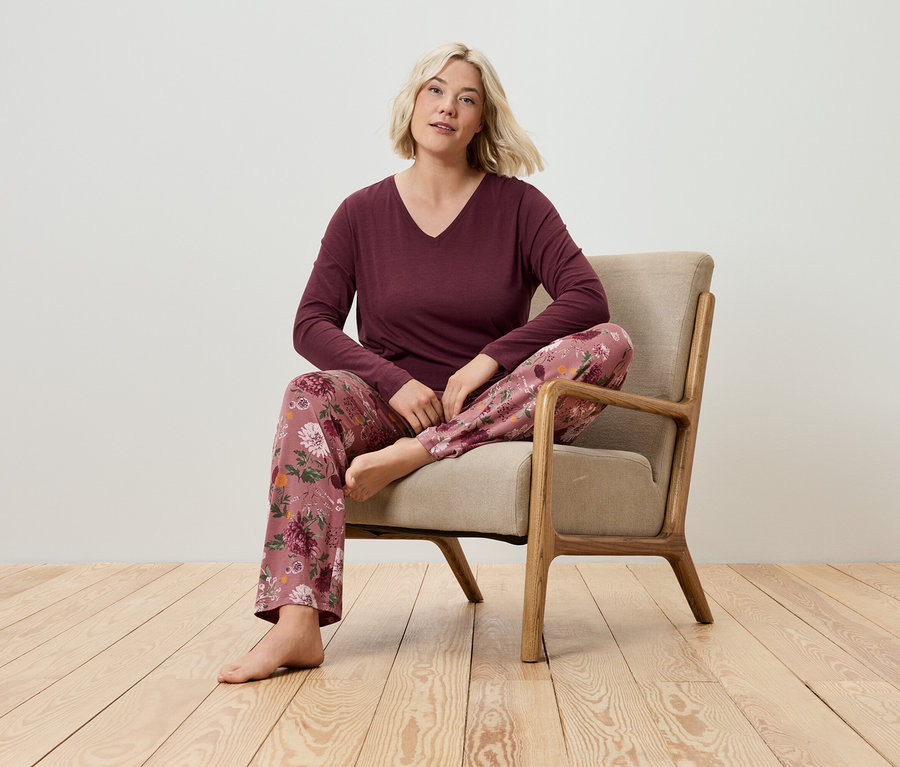 Blonde Frau sitzt in einem Sessel und trägt ein burgunderrotes Langarmshirt und eine Schlafanzughose mit Blumenmuster.