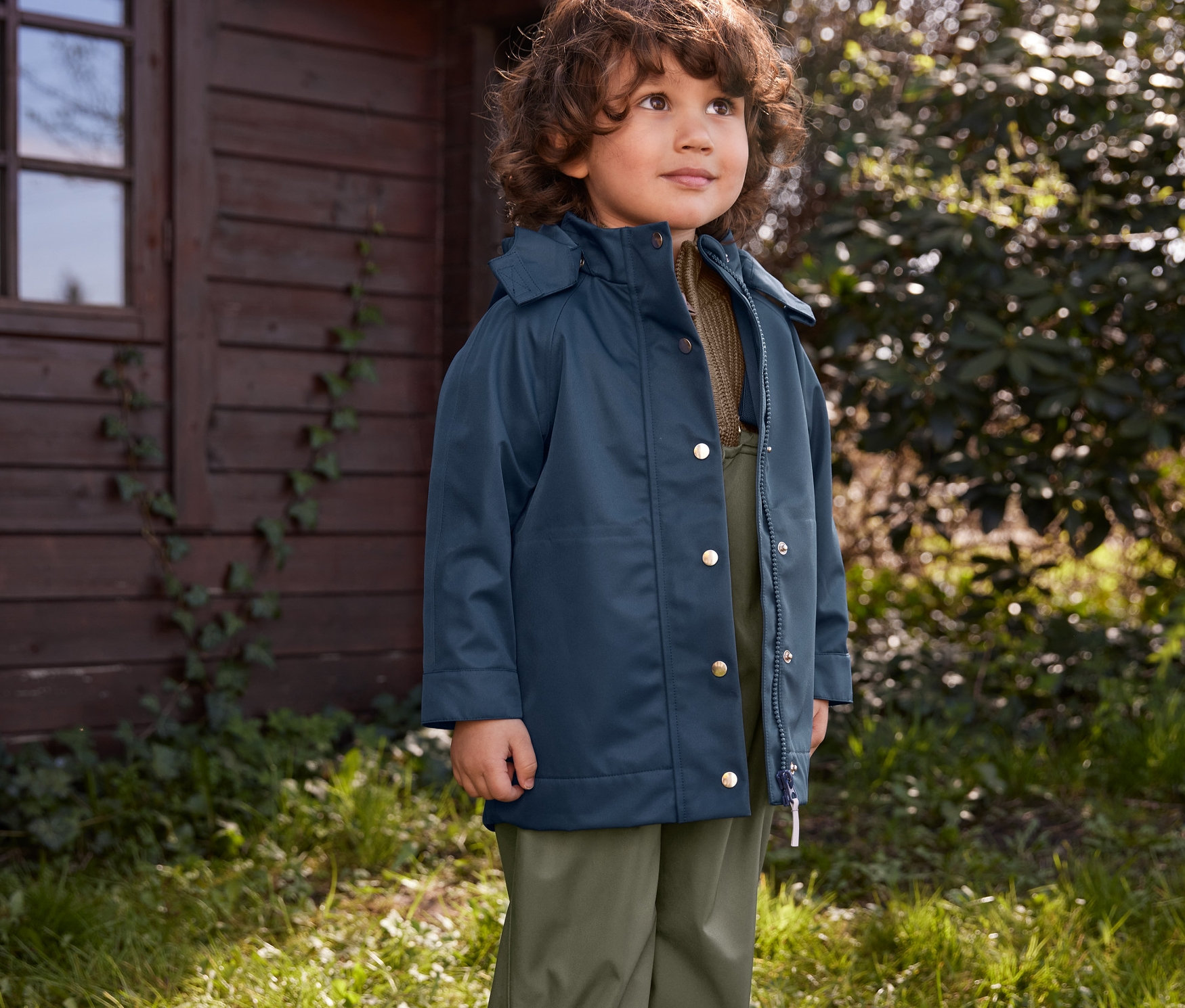 Ein Junge steht draußen im Gras und trägt Gummistiefel, eine Regenhose und eine Regenjacke.