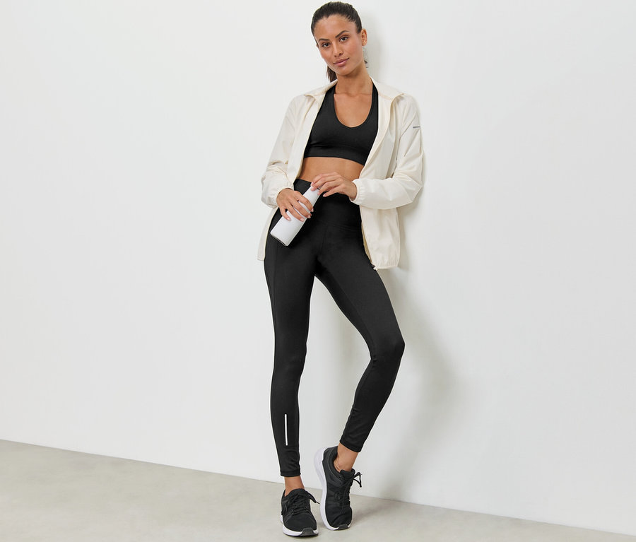 Eine Frau lehnt an einer Wand, hält eine Wasserflasche und trägt einen Sport-BH, eine Jacke und eine wasserabweisende Sporttight.