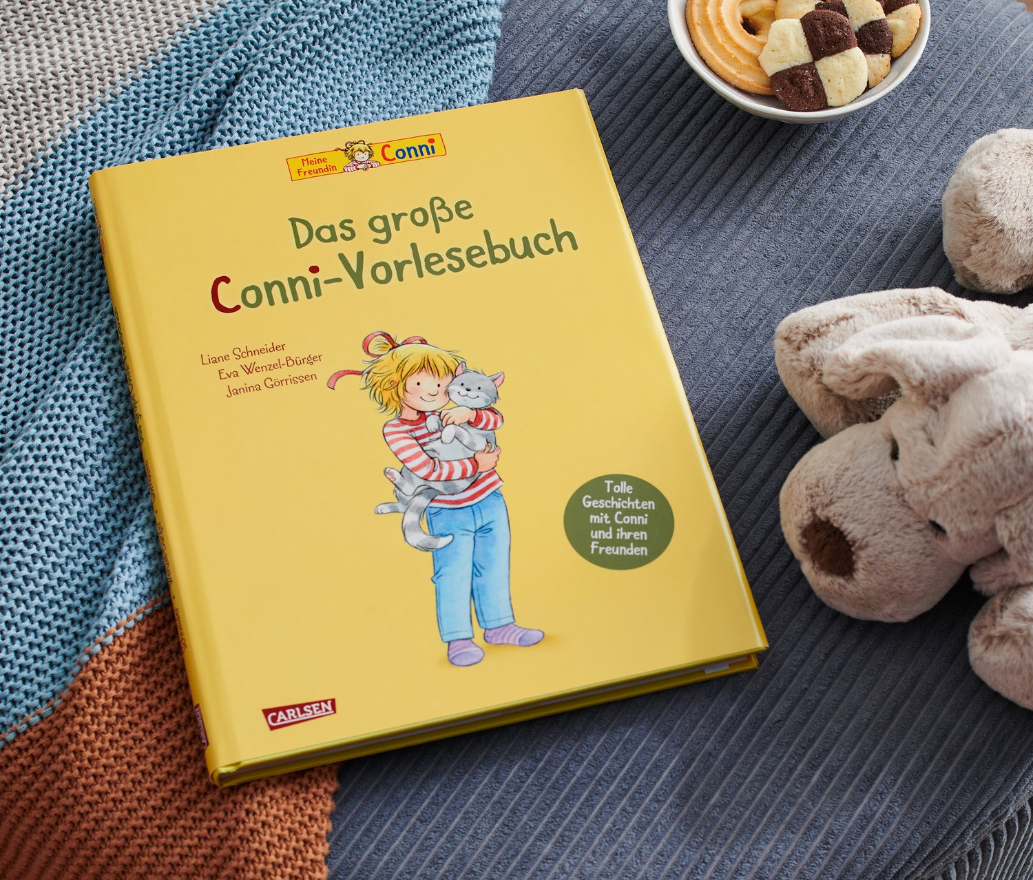 Auf einem blauen Hocker liegen das gelbe Buch »Das große Conni-Vorlesebuch«, ein Plüschhund, eine Schale Kekse und eine Strickdecke.