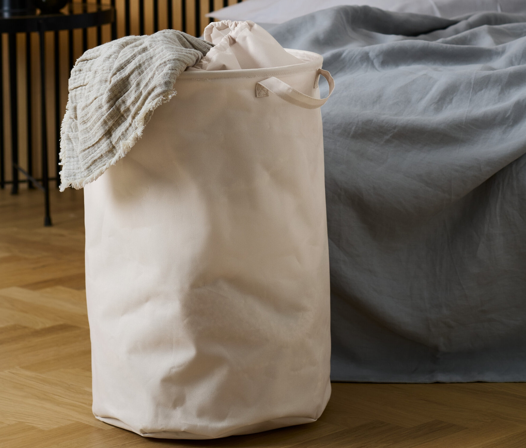 Ein beiger Wäschesammler mit einer beigen Decke, die darüber hängt, steht neben einem Bett.