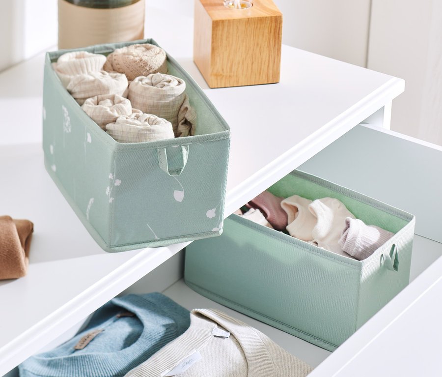 Zwei mittelgroße Schubladen-Ordnungsboxen auf einer weißen Kommode. Eine Box befindet sich auf der Oberseite der Kommode und enthält zusammengerollte Kleidung. Die zweite befindet sich in einer offenen Schublade mit Kleidung.