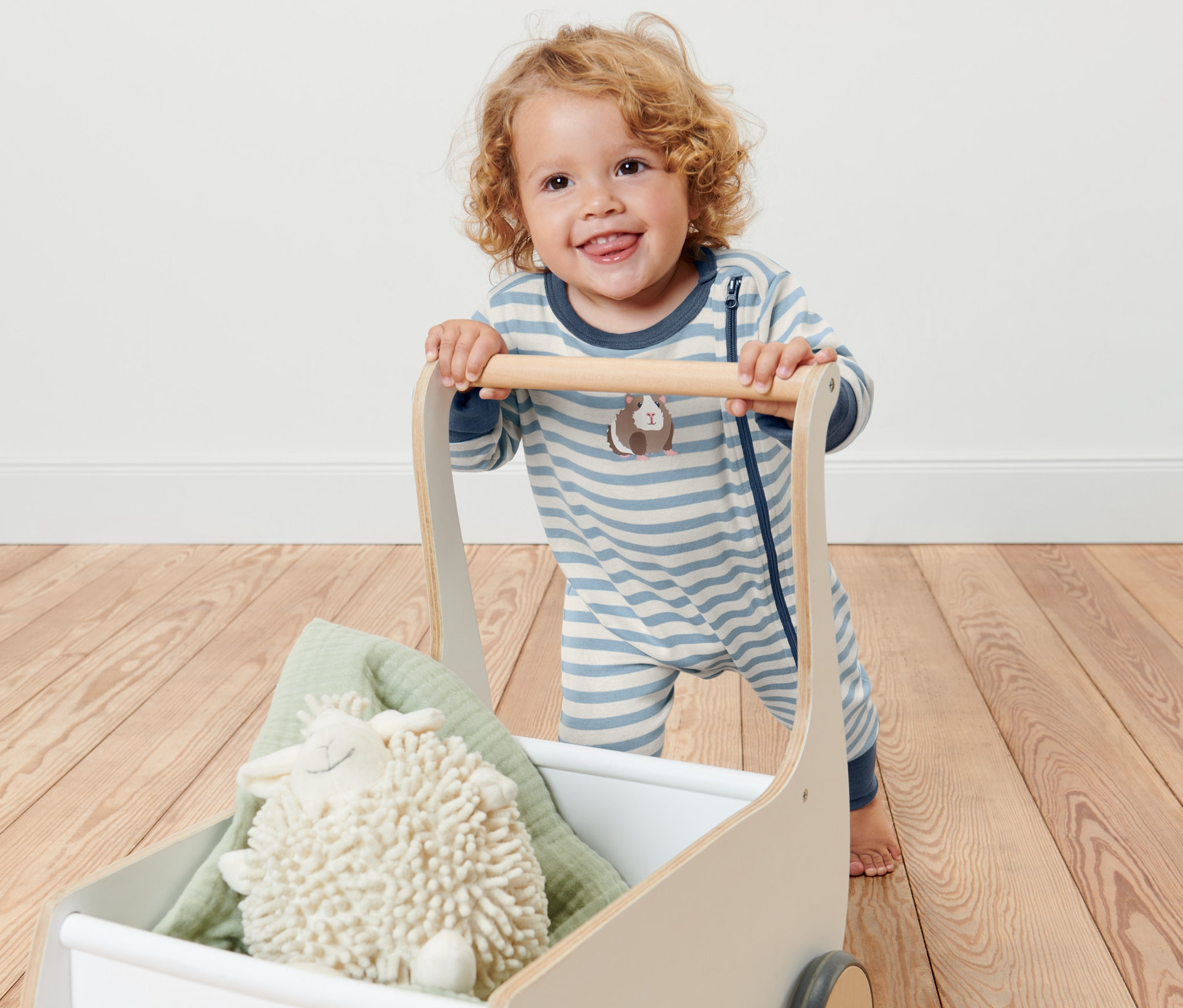 Ein lächelndes Kind schiebt einen Wagen mit einem gestreiften Baby-Pyjama, einem Plüschschaf und einer grünen Decke.