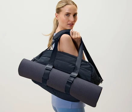 Blonde Frau trägt eine schwarze Sporttasche und eine Yogamatte über der Schulter.