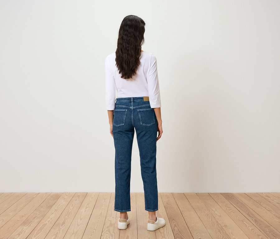 Eine Frau mit langen, dunklen Haaren ist von hinten in blauen Jeans und einem weißen T-Shirt zu sehen.