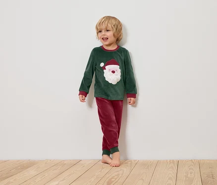 Ein Junge im grünen Weihnachtspyjama mit Santa-Claus-Motiv steht auf einem Holzboden vor einer weißen Wand.