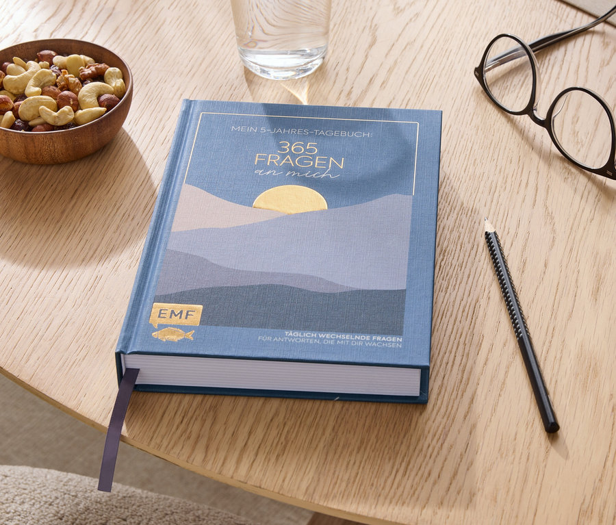 Auf einem Holztisch liegen ein Buch, ein Glas Wasser, eine Schale Nüsse, eine Brille und ein Stift.