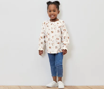 Lächelndes kleines Mädchen posiert im Studio und trägt eine Kinder-Musselin-Bluse, Jeans und Turnschuhe.