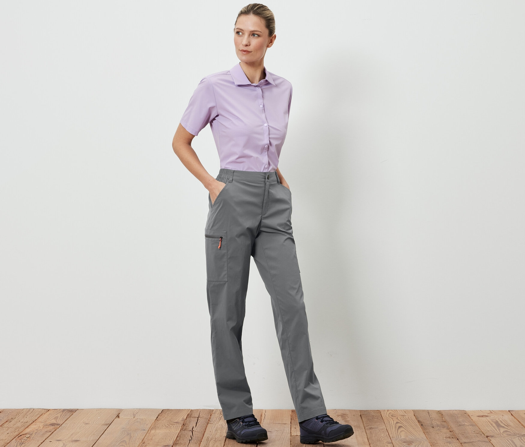 Eine Frau steht und posiert in grauen, packbaren Outdoorhosen und einem hellvioletten Hemd.