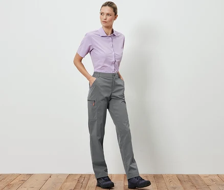 Eine Frau steht und posiert in grauen, packbaren Outdoorhosen und einem hellvioletten Hemd.