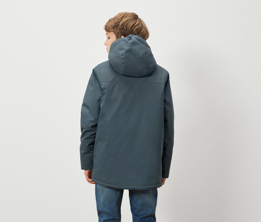 Ein Junge in einer blauen Kapuzenjacke und blauen Jeans steht mit dem Rücken zur Kamera vor einem weißen Hintergrund.