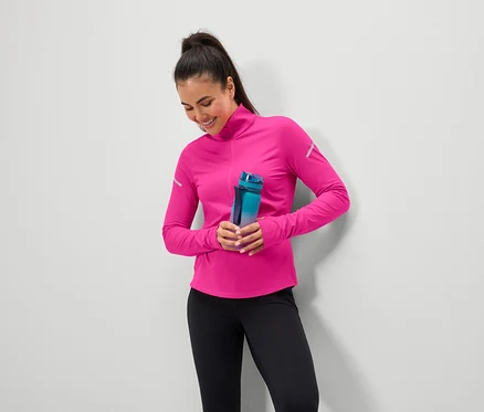 Lächelnde Frau hält eine blaugrüne Wasserflasche, trägt ein pinkes Oberteil und schwarze Leggings.