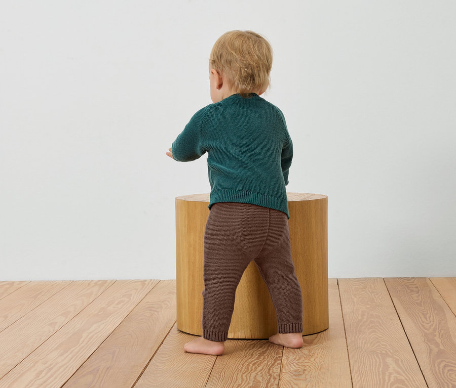 Kleinkind in grünem Pullover und braunen Leggings steht an einem Holzhocker.