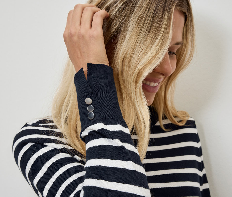 Blonde Frau streicht sich eine Haarsträhne aus dem Gesicht. Sie trägt einen gestreiften Pullover mit Knöpfen am Ärmel.