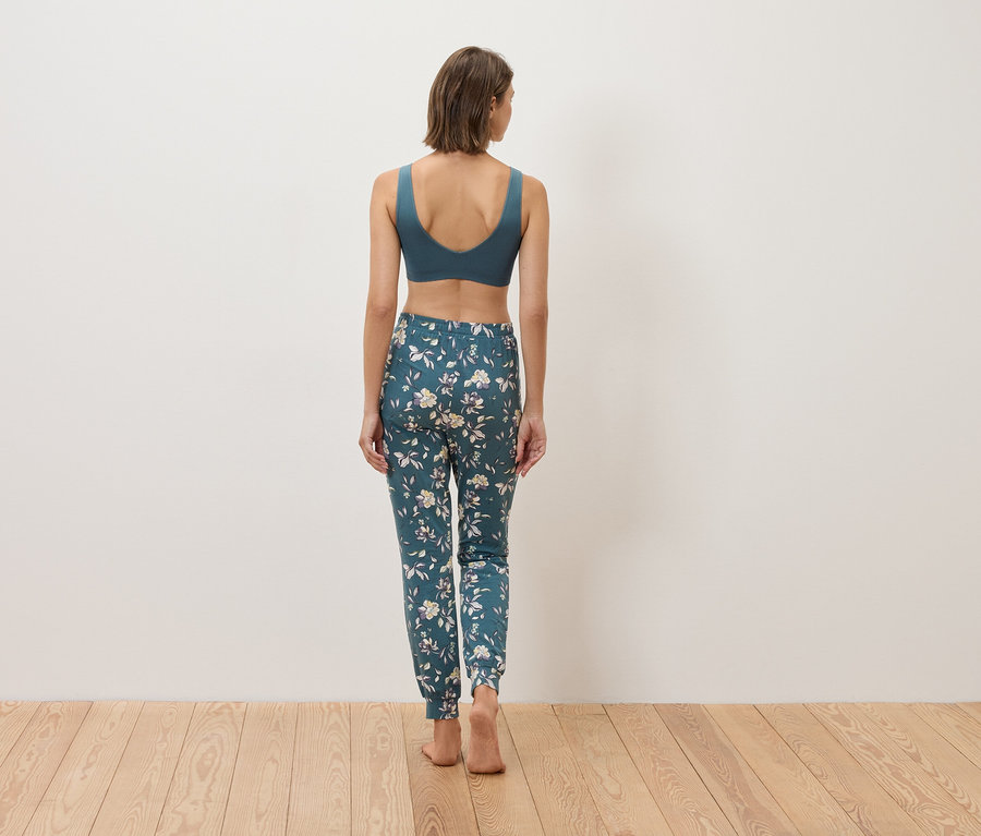 Rückansicht einer Frau mit blauem Sport-BH und blumiger Hose auf Holzboden.