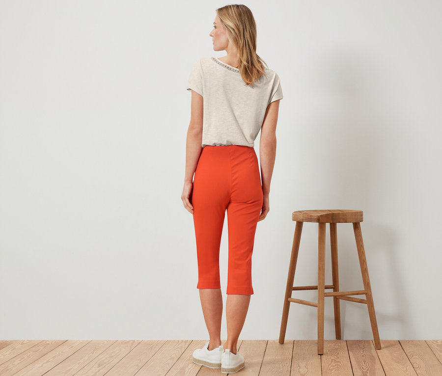 Frau steht von hinten, trägt eine orangefarbene 3/4-Stretchhose.