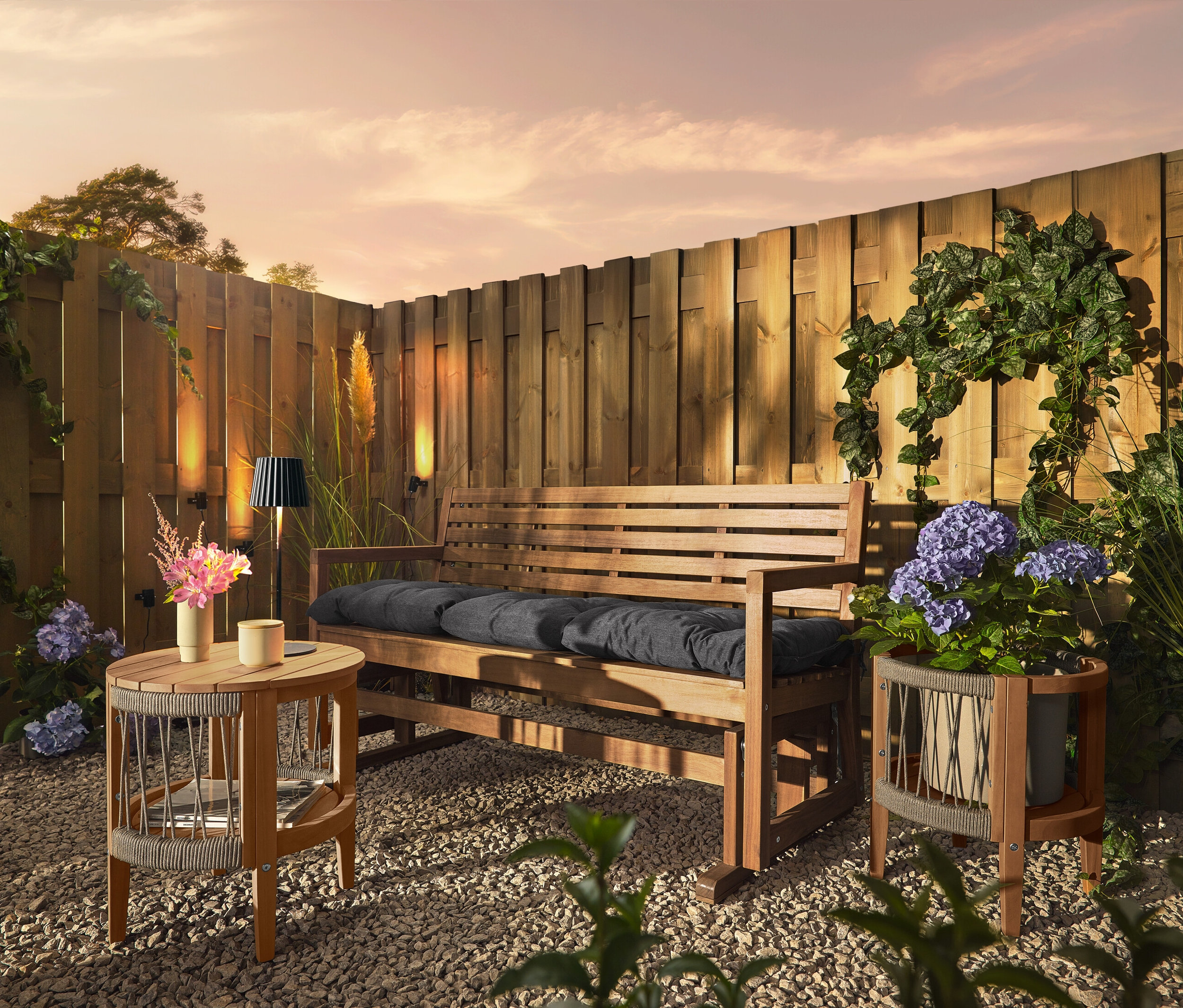 Eine Holzbank mit Kissen steht auf einem Kiesboden vor einem Holzzaun. An dem Zaun sind drei Solar-Dachrinnenleuchten befestigt.
