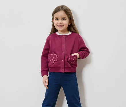 Lächelndes Mädchen mit braunen Haaren, trägt eine weinrote Strickjacke mit blumenförmigen Taschen und blaue Hosen vor einem weißen Hintergrund.