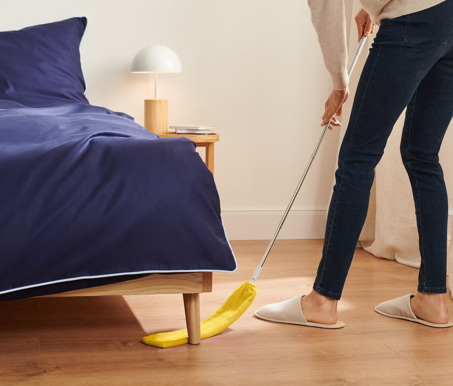 Eine Frau wischt mit einem gelben Staubwedel unter einem Bett mit marineblauer Bettwäsche.
