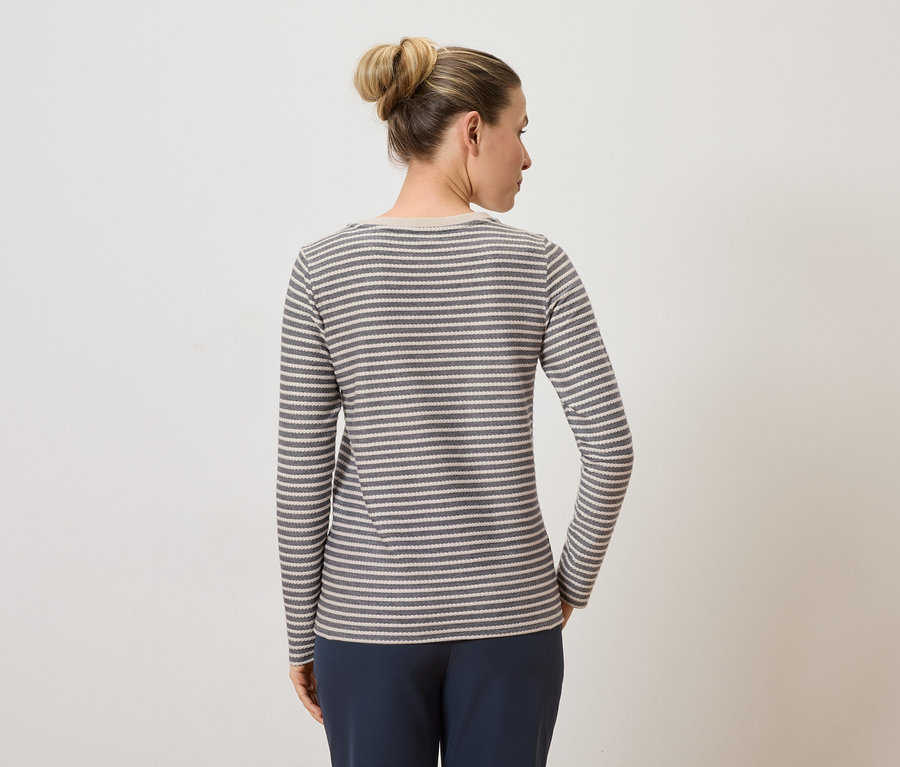 Frau mit Duttfrisur, von hinten, trägt ein grau-weiß gestreiftes Langarmshirt und blaue Hose vor einem hellen Hintergrund.