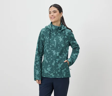 Frau mit Pferdeschwanz trägt eine grüne, blumengemusterte Jacke mit Kapuze und blaue Hose vor einem weißen Hintergrund.