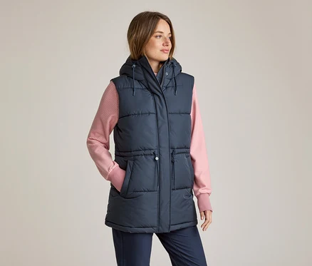 Frau in einer marineblauen, wattierten Kapuzenweste über einem rosa-weiß gestreiften Langarmshirt und marineblauen Hosen, die linke Hand in der Tasche, blickt nach rechts.