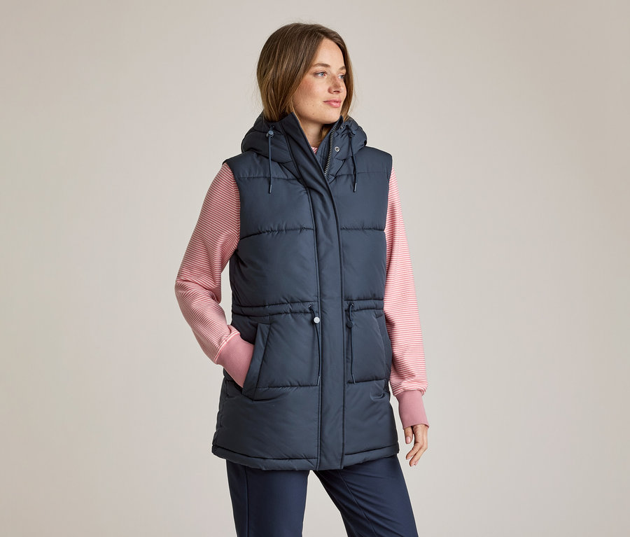 Frau in einer marineblauen, wattierten Kapuzenweste über einem rosa-weiß gestreiften Langarmshirt und marineblauen Hosen, die linke Hand in der Tasche, blickt nach rechts.