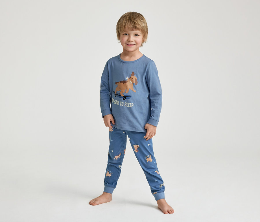 Ein lächelnder kleiner Junge steht barfuß in einem blauen Schlafanzug mit einem Hund auf einem Skateboard-Aufdruck und der Aufschrift "TOO COOL TO SLEEP".