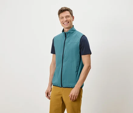 Mann mit blaugrüner Weste, marineblauem T-Shirt und ockerfarbener Hose lächelt vor einem weißen Hintergrund.