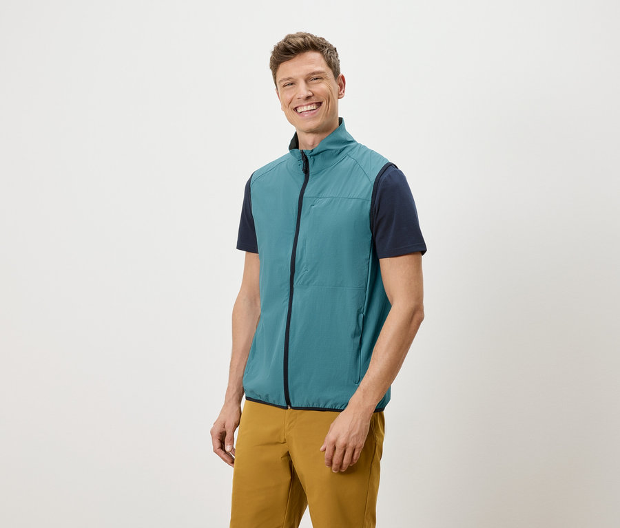 Mann mit blaugrüner Weste, marineblauem T-Shirt und ockerfarbener Hose lächelt vor einem weißen Hintergrund.
