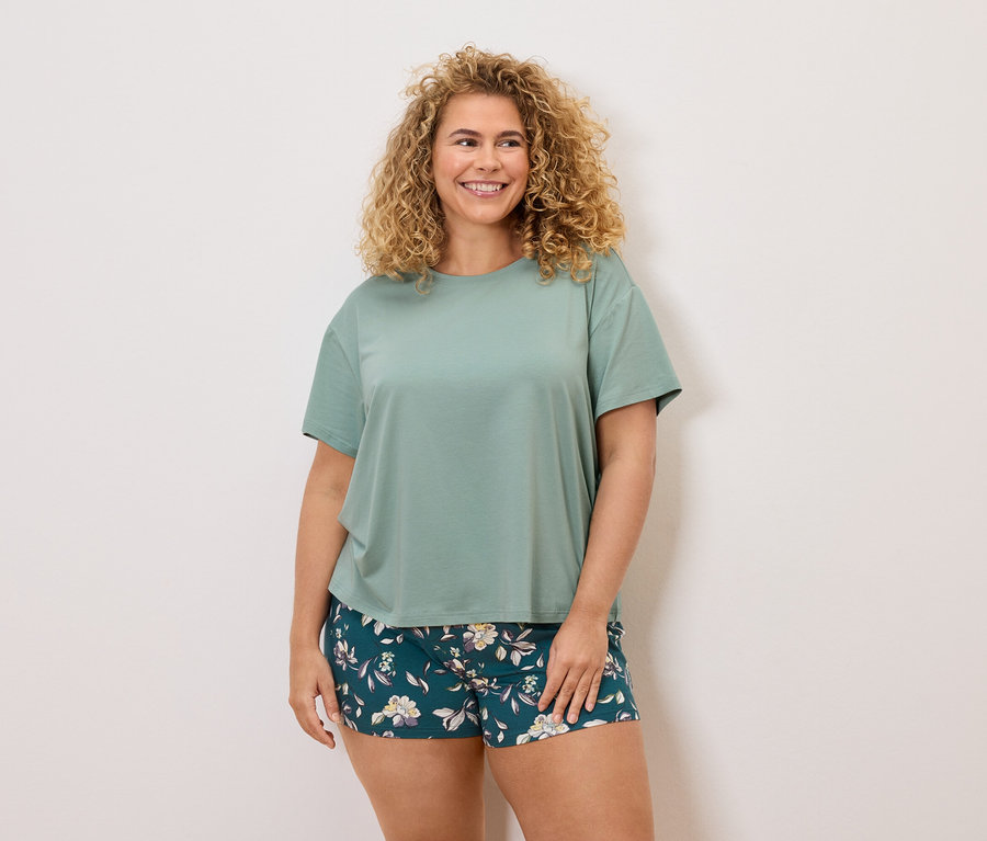 Frau mit lockigem Haar, trägt ein grünes T-Shirt und Shorts mit Blumenmuster.