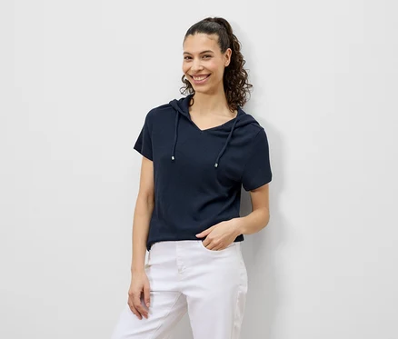 Frau mit lockigem Haar lächelt in marineblauem Kapuzen-T-Shirt und weißen Hosen.