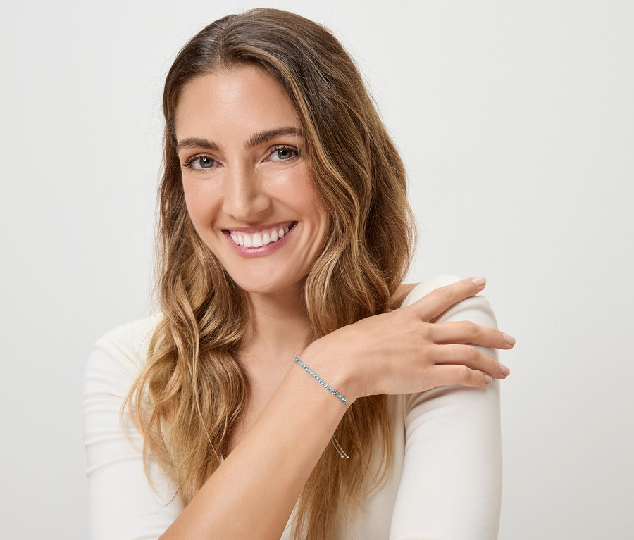 Lächelnde Frau mit blonden, gewellten Haaren und blauem Armband am linken Handgelenk vor weißem Hintergrund.