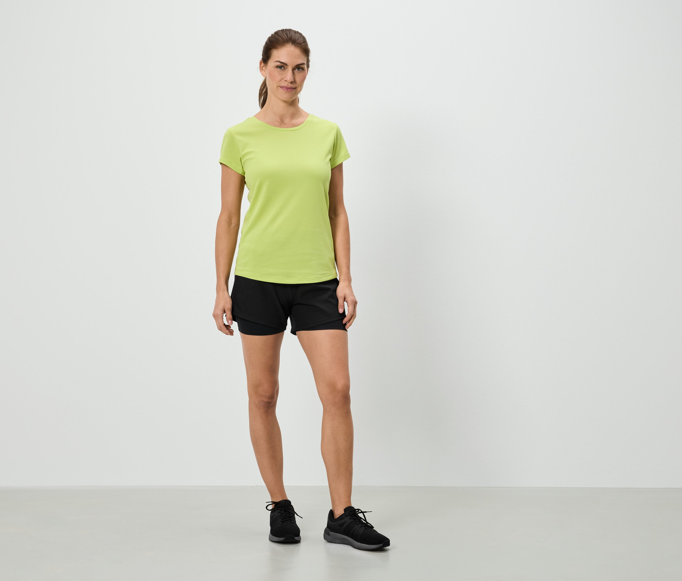 Eine Frau steht in einem hellgrünen T-Shirt, schwarzen Shorts und schwarzen Turnschuhen vor einer weißen Wand.