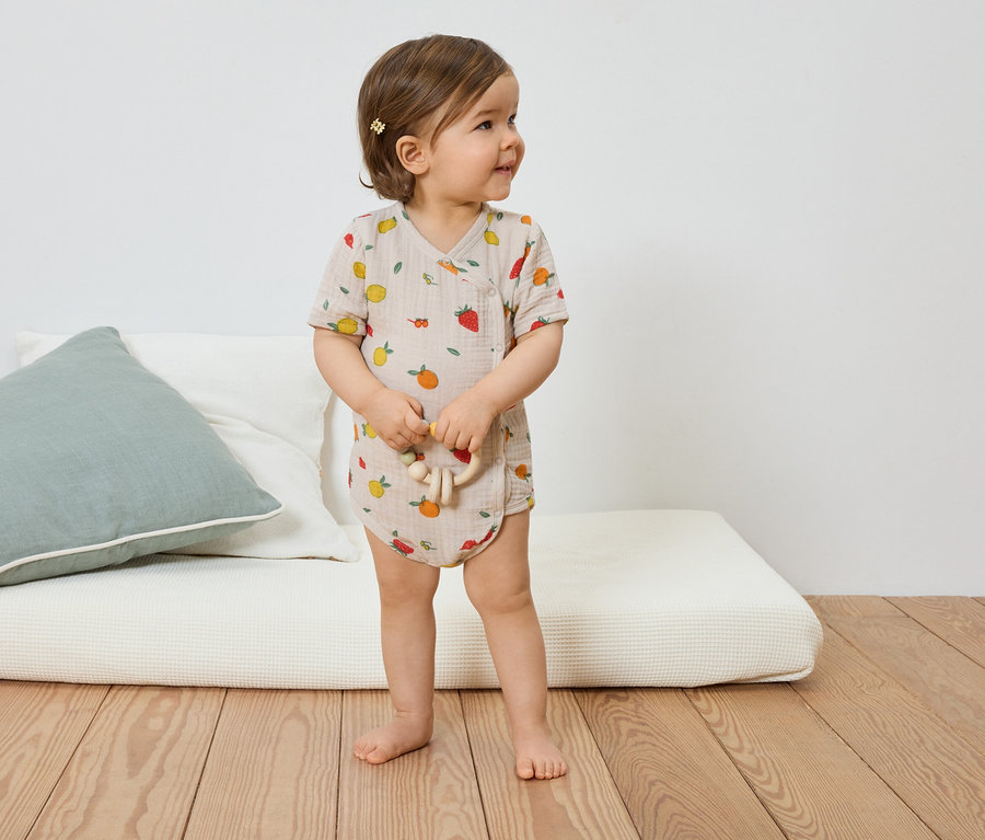 Ein Kleinkind mit Haarspange trägt einen hellbraunen Kurzarmbody mit Fruchtmuster und hält ein Holzspielzeug in der Hand.