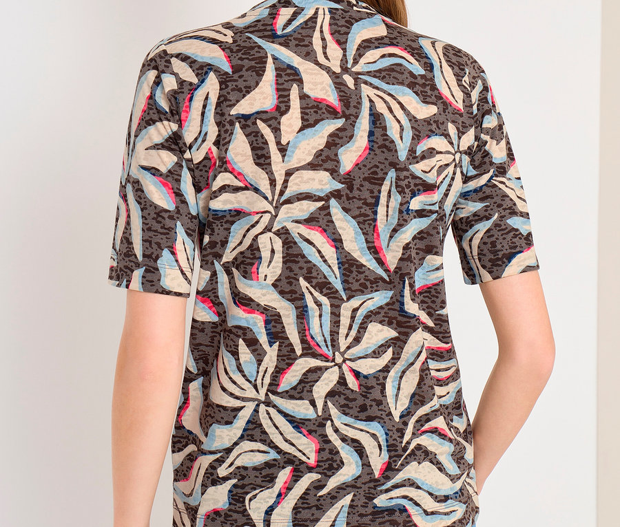 Frau von hinten in einem dunkel gemusterten CECIL Kurzarmshirt mit hellen, blauen und pinken Blattformen und blauen Jeans.