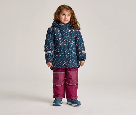 Ein kleines Mädchen mit lockigem Haar steht in einer blauen, geblümten Jacke und einer weinroten Hose.