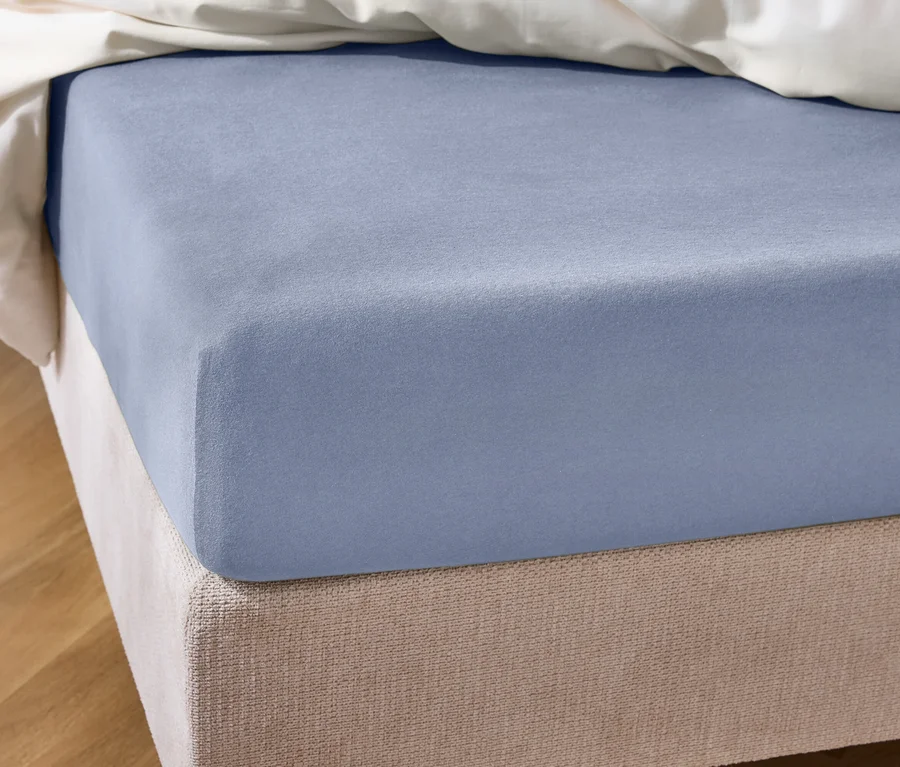 Nahaufnahme eines Bettes mit hellblauem Spannbettlaken und beiger Bettumrandung unter einer weißen Decke.