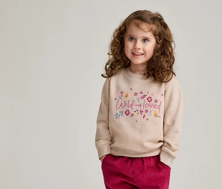 Mädchen mit lockigem Haar trägt einen hellbraunen Pullover mit der Aufschrift "Wild and loved" und eine rote Hose.