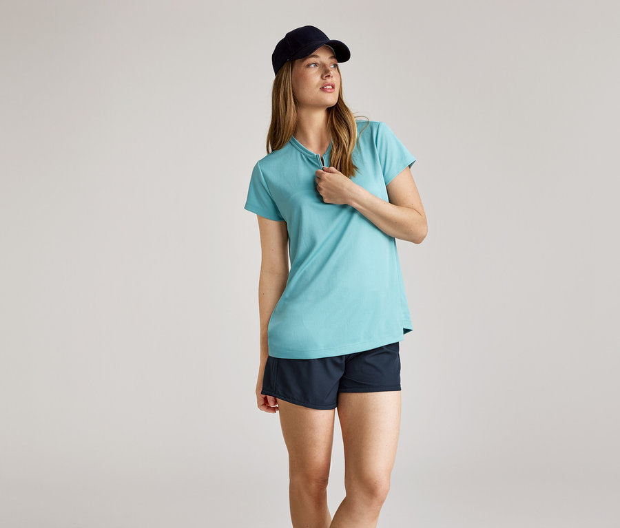 Frau mit dunkelblauer Kappe, hellblauem T-Shirt mit kurzem Reißverschluss und dunkelblauen Shorts vor hellem Hintergrund.