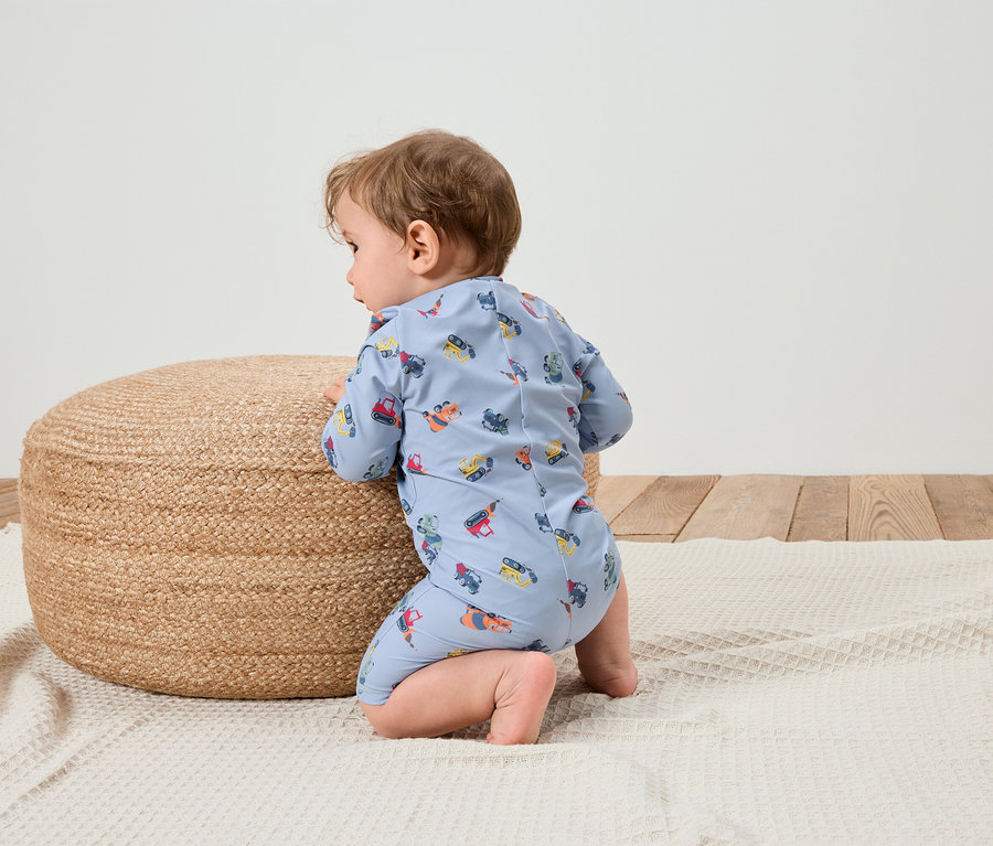 Baby in hellblauem Overall mit Baufahrzeugmuster kniet auf einer weißen Decke neben einem runden, geflochtenen Pouf.