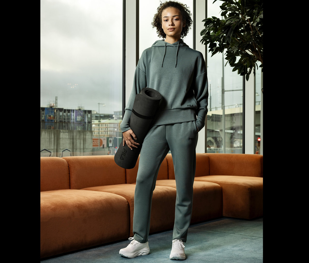 Frau im grünen Trainingsanzug hält eine Yogamatte vor einem Fenster.