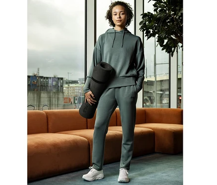 Frau in salbeigrüner FILA Modalmix-Jogginghose und Kapuzenpullover, hält eine schwarze Yogamatte, steht vor einem Fenster mit Stadtblick.