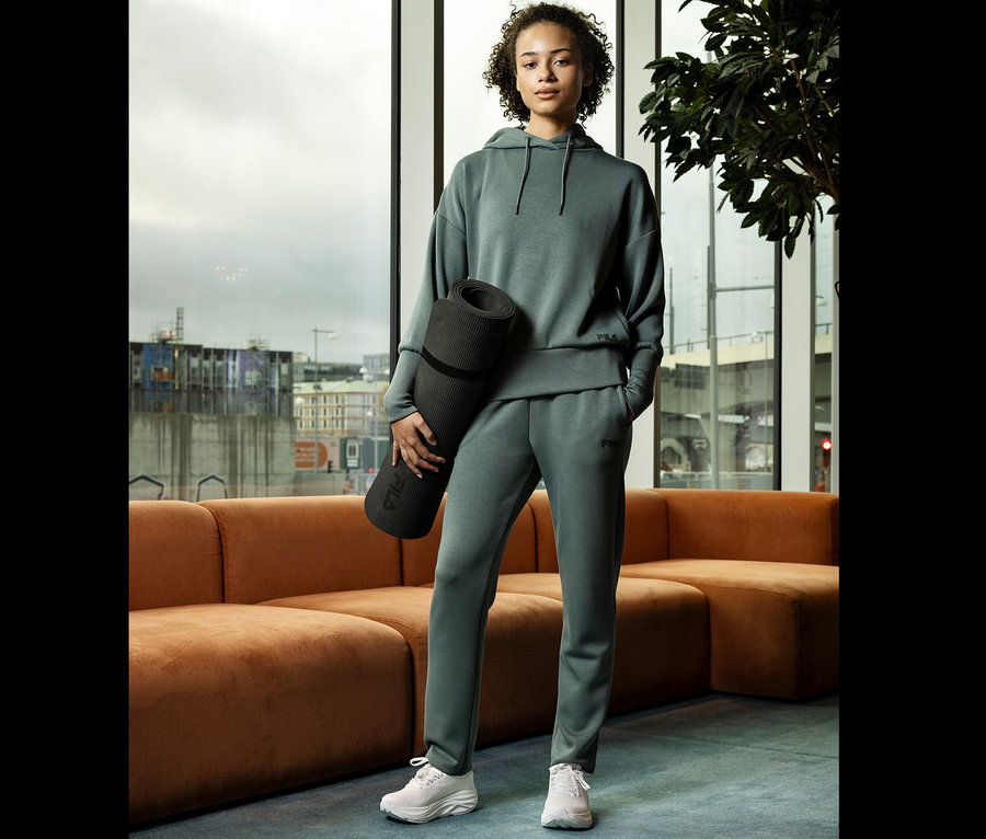 Frau im grünen Trainingsanzug hält eine Yogamatte vor einem Fenster.