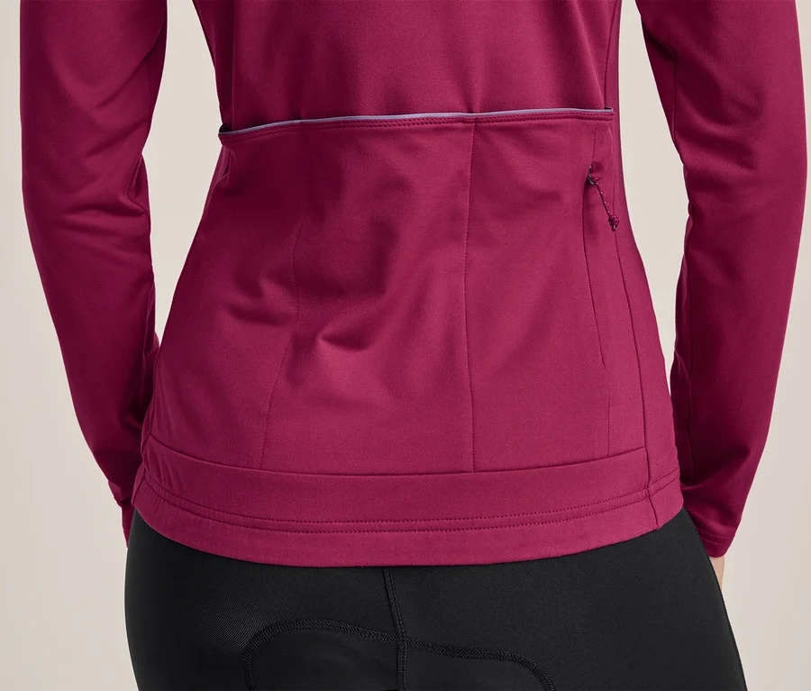 Person von hinten in einem langärmeligen, magentafarbenen Radtrikot und schwarzen Radhosen. Das Trikot hat drei Rückentaschen, eine davon mit Reißverschluss, und einen reflektierenden Streifen.